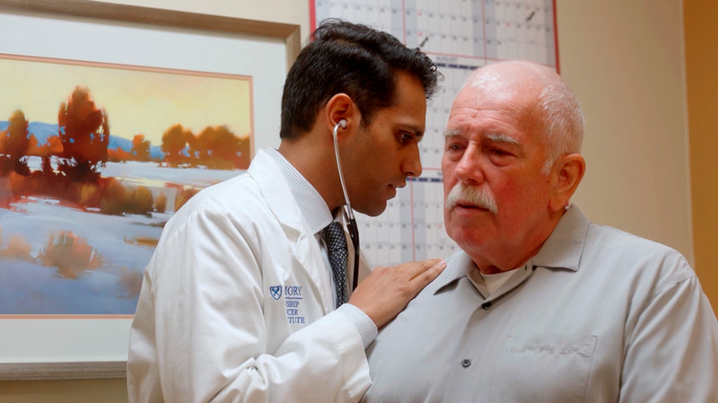 Man sits in an exam room while a physician places a stethoscope on his back.