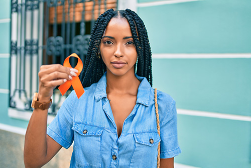 Photo: Young African American woman looking into camera lense and holding orange cancer ribbon