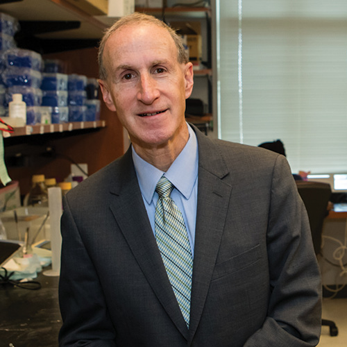 a portrait of an older white researcher standing in his lab and smiling to the camera.