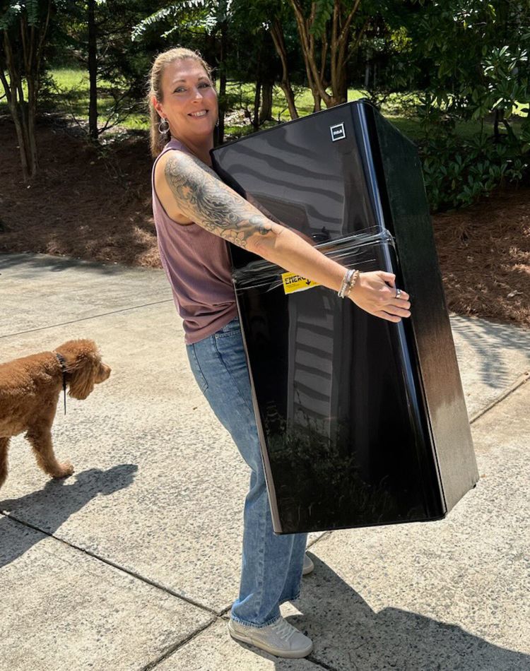 a photography of a female lifting a small fridge in the driveway.